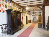 Ein historischer Flur mit Holzbalken, Wappen an den Wänden und einem großen, verzierten Schrank neben einem roten Teppich.