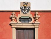 Ein rotes Schloss mit verzierten Steinskulpturen und einem kunstvollen Wappen über der Eingangstür.
