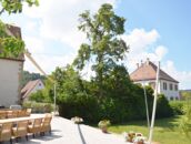 Ein sonniger Garten mit einem langen Tisch und Stühlen neben einem großen Baum und zwei traditionellen Häusern.
