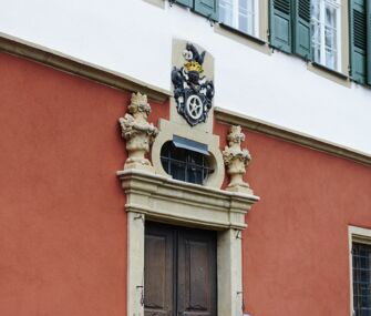 Eine historische Tür mit kunstvollen Verzierungen und einem Wappen ziert die rote Fassade eines alten Gebäudes.