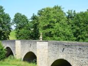 Eine alte Steinbrücke mit drei Bögen führt durch eine grüne Landschaft, während eine Statue darauf steht.