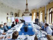 In einem eleganten Raum des Schlosses nehmen Personen an einem Pressegespräch teil, während Präsentationen vorbereitet werden.