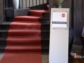 Ein roter Teppich führt eine Treppe hinauf, flankiert von Blumen, während ein Schild auf Schulungsräume hinweist.