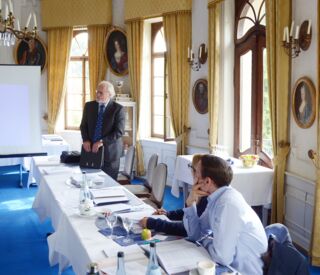 Ein Redner präsentiert bei einem Pressegespräch auf einem eleganten Schlosscampus vor aufmerksamem Publikum.