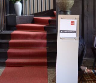 Ein roter Teppich führt eine Treppe hinauf, flankiert von Pflanzen, mit einem Schild zu Schulungsräumen im Schlosscampus.