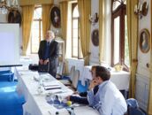 In einem eleganten Raum des Schlosses hält ein Mann ein Pressegespräch mit mehreren aufmerksam zuhörenden Teilnehmern.