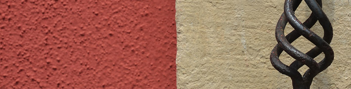 Ein kunstvolles Metallgitter hebt sich vor einer rot-beige geteilten Wand ab.